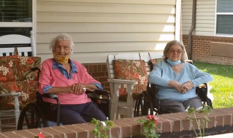 Two elderly women sitting in wheelchairs on a covered patio outside a building. Both women are wearing face masks pulled down below their chins. There are floral patterned cushions on white wooden chairs behind them and a small garden with flowers in front of a low brick wall.