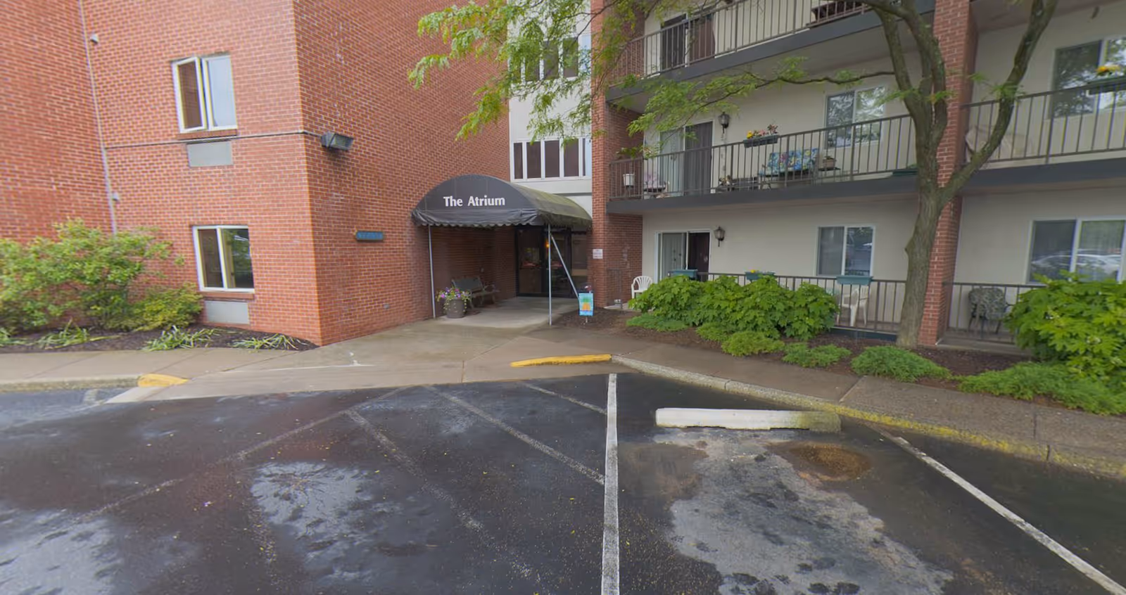 Exterior view of a senior living facility entrance named The Atrium, showing a brick building with balconies, some greenery, and a wet parking lot in front.