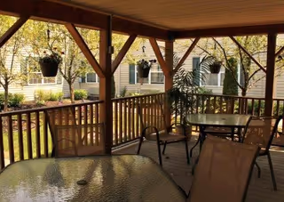Covered outdoor patio area with glass-top tables and chairs, hanging flower pots, and a view of trees and a building with windows in the background.