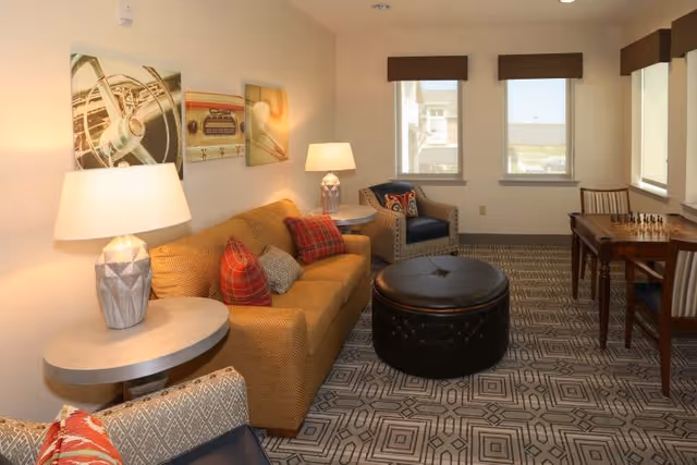 A cozy living room with a mustard yellow sofa adorned with red and gray plaid pillows, a round black leather ottoman in the center, two patterned armchairs, a side table with a lamp, and a wooden table with a chessboard near three windows with brown valances.