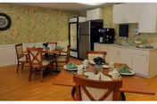 A dining area with wooden floors and floral wallpaper. There are two wooden tables set with green napkins, white cups, and plates. Four wooden chairs with cushioned seats surround each table. In the background, there is a kitchenette area with white cabinets, a black refrigerator, a coffee machine, and a paper towel dispenser.