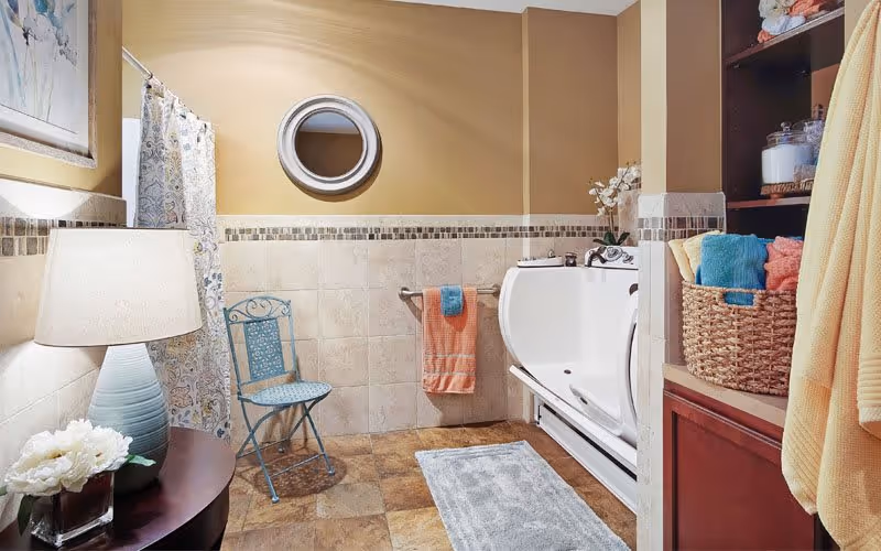 A bathroom with beige walls and tiled floor featuring a walk-in bathtub with grab bars, a blue metal chair, a round mirror above the bathtub, a towel rack with orange and blue towels, a wooden shelf with baskets and towels, a table with a lamp and a flower vase, and a patterned shower curtain.