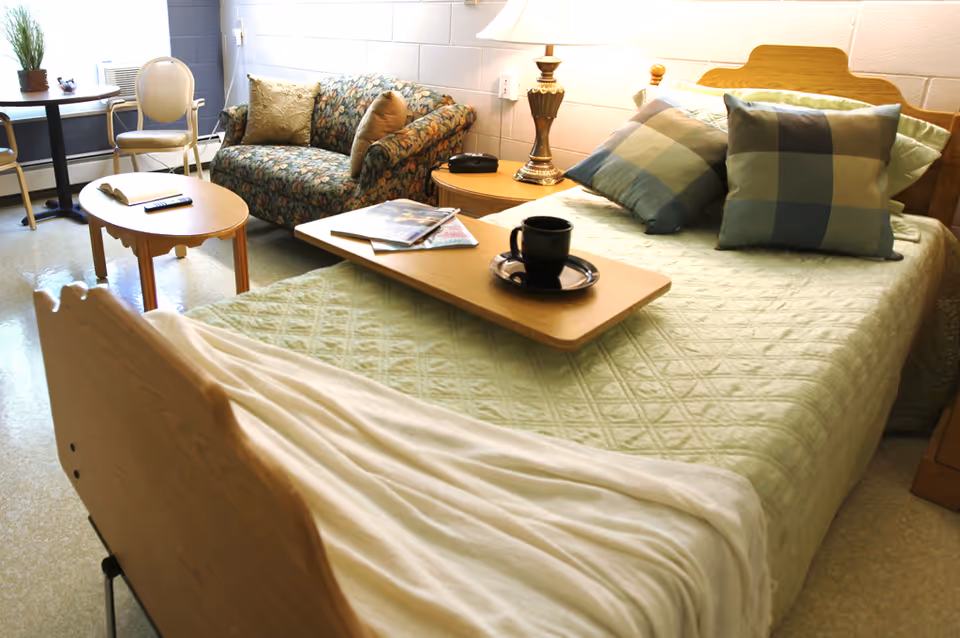 A cozy bedroom in a senior living facility featuring a bed with a light green quilt, plaid pillows, and a wooden headboard. A wooden tray with a black cup and saucer, along with magazines, rests on the bed. Next to the bed is a wooden nightstand with a lamp and a phone. In the background, there is a floral-patterned loveseat, a small wooden coffee table with a remote control and book, and a chair near a window with a small potted plant on the windowsill.