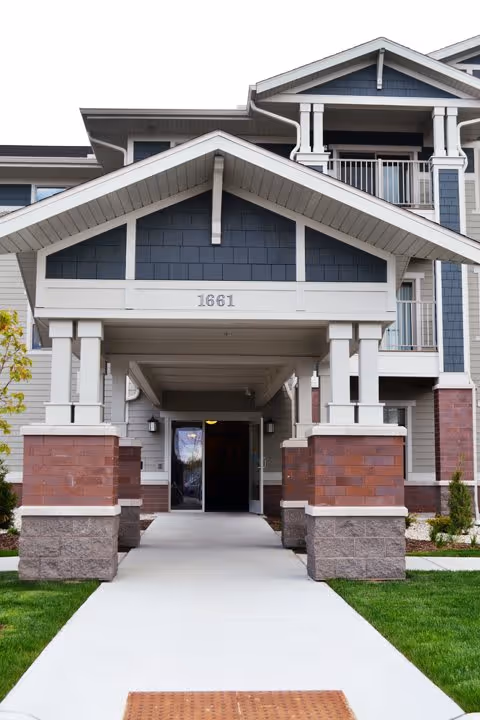 Entrance of a multi-story senior living facility with a covered porch supported by white pillars and brick bases, the building number 1661 displayed above the entrance, and a concrete walkway leading up to the door.