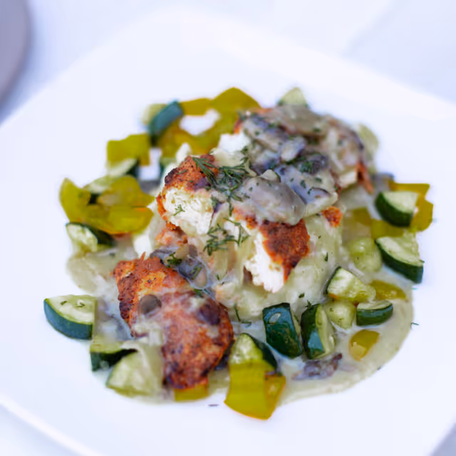 A plated dish featuring grilled or roasted chicken pieces topped with a creamy mushroom sauce, served on a bed of mashed potatoes and garnished with chopped green vegetables including zucchini and bell peppers.