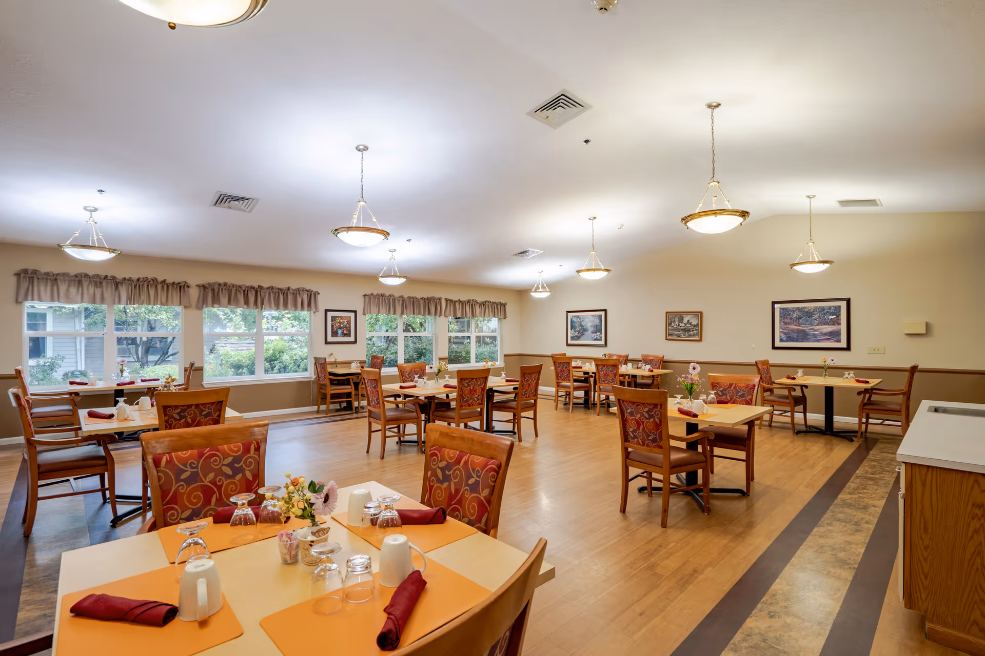 A spacious dining room with multiple wooden tables and chairs arranged neatly. Each table is set with placemats, napkins, glasses, and small flower arrangements. Large windows with valance curtains allow natural light to brighten the room. The walls are decorated with framed artwork, and ceiling lights provide additional illumination.