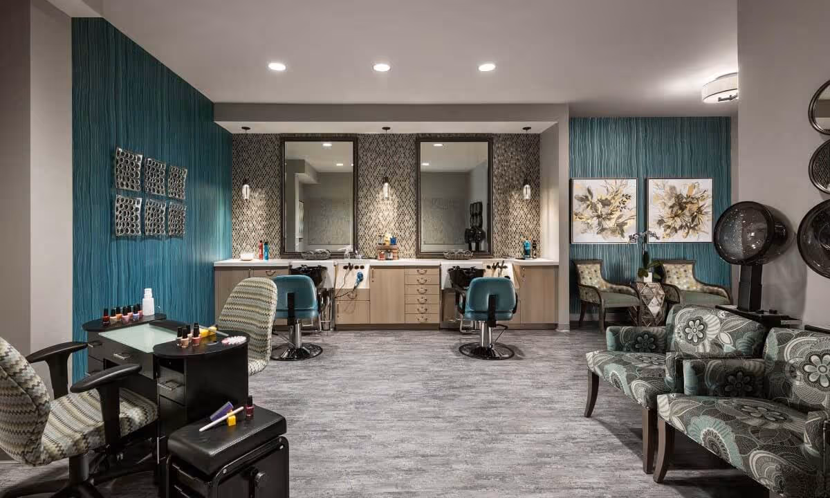 A stylish salon area in a senior living facility with two teal salon chairs in front of large mirrors and wash basins. To the left, there is a manicure station with nail polish bottles and two patterned chairs. On the right, there are patterned armchairs and two hair dryers. The walls have teal textured wallpaper and decorative artwork.