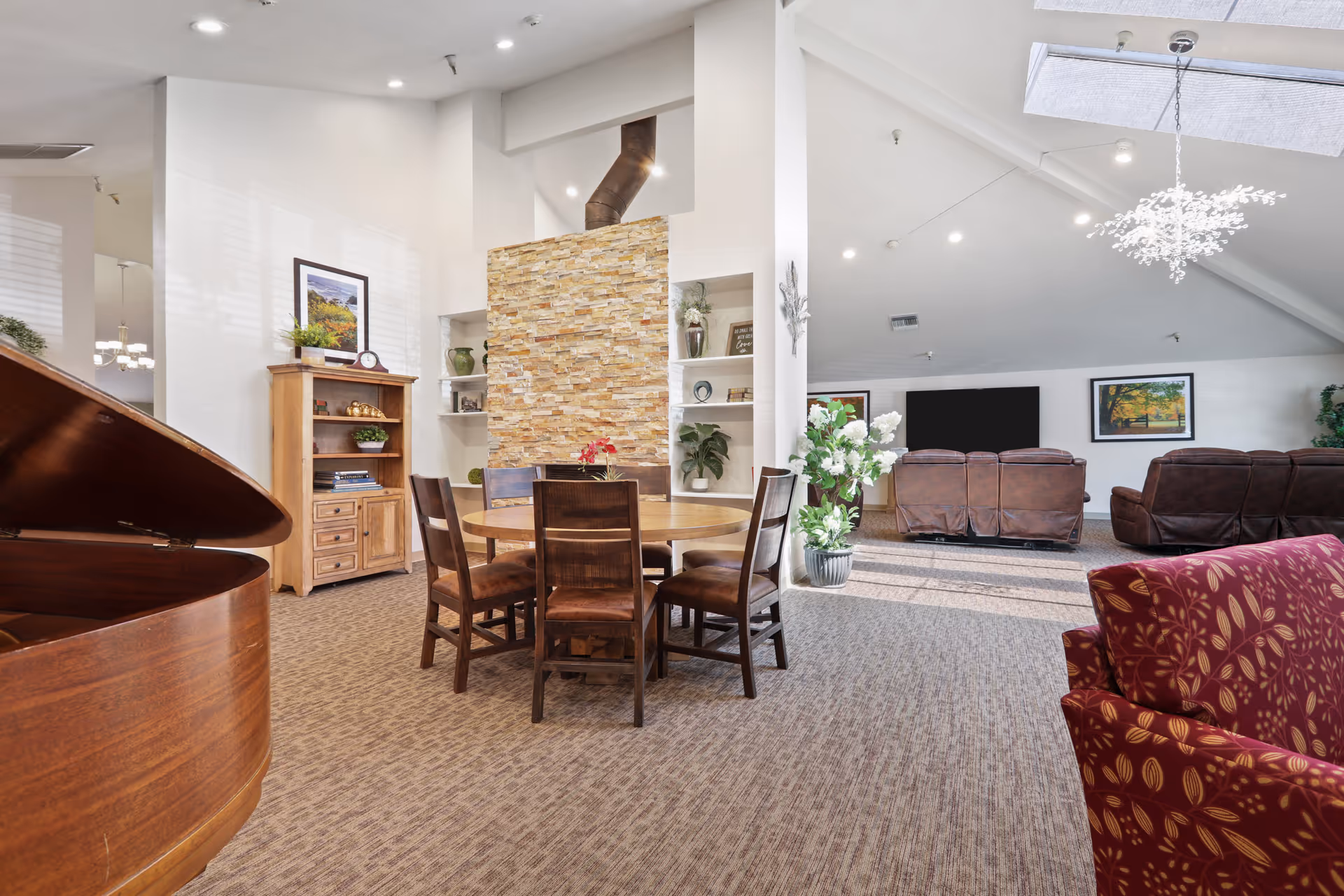 A bright and spacious assisted living common area featuring a round wooden table with six chairs, a stone accent wall with a chimney, built-in shelves with decorative items, a wooden cabinet with a framed picture and plants, a red patterned armchair, and a seating area with brown leather recliners facing a large flat-screen TV. The room has a high ceiling with recessed lighting and a skylight, and a modern chandelier hangs from the ceiling.