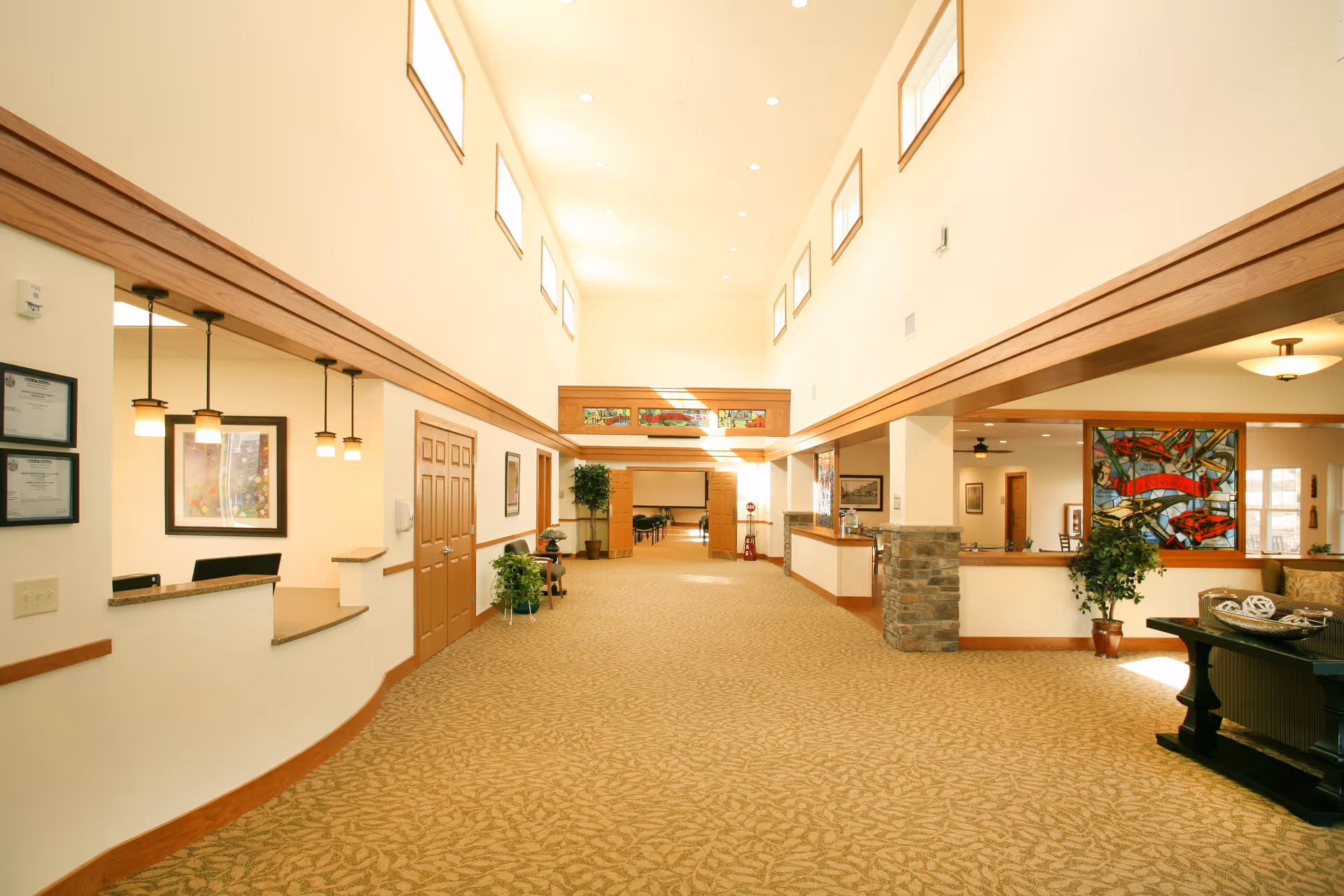 Bright and spacious hallway in a senior living facility with high ceilings and multiple windows near the ceiling allowing natural light. The hallway features beige patterned carpet, wooden trim, and light-colored walls. On the left side, there is a reception area with hanging pendant lights and framed certificates on the wall. On the right side, there is a seating area with a table, plants, and a colorful stained glass window. The hallway leads to another room with tables and chairs visible in the distance.
