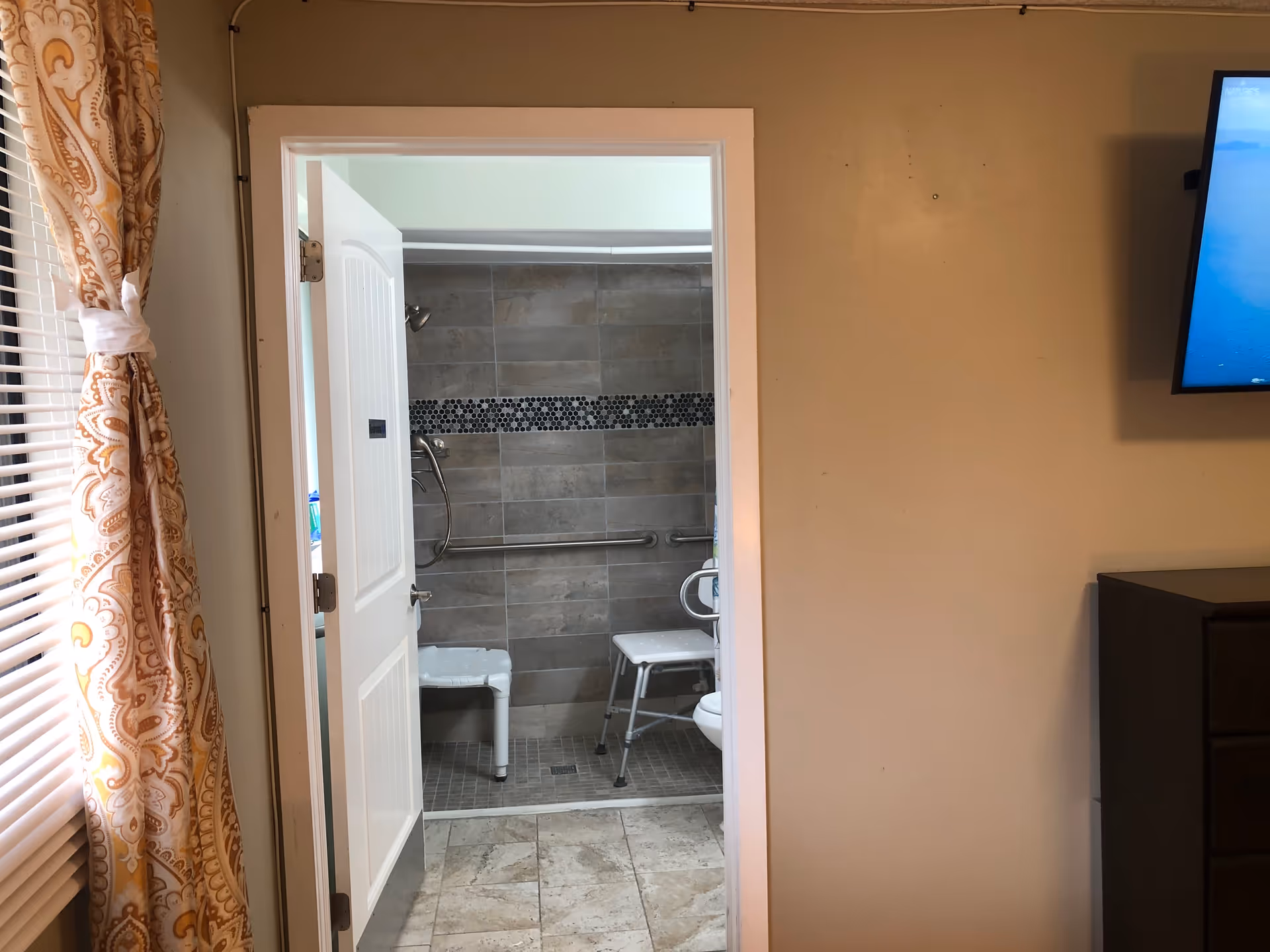 Open doorway revealing an accessible tiled bathroom with grab bars and shower chairs.