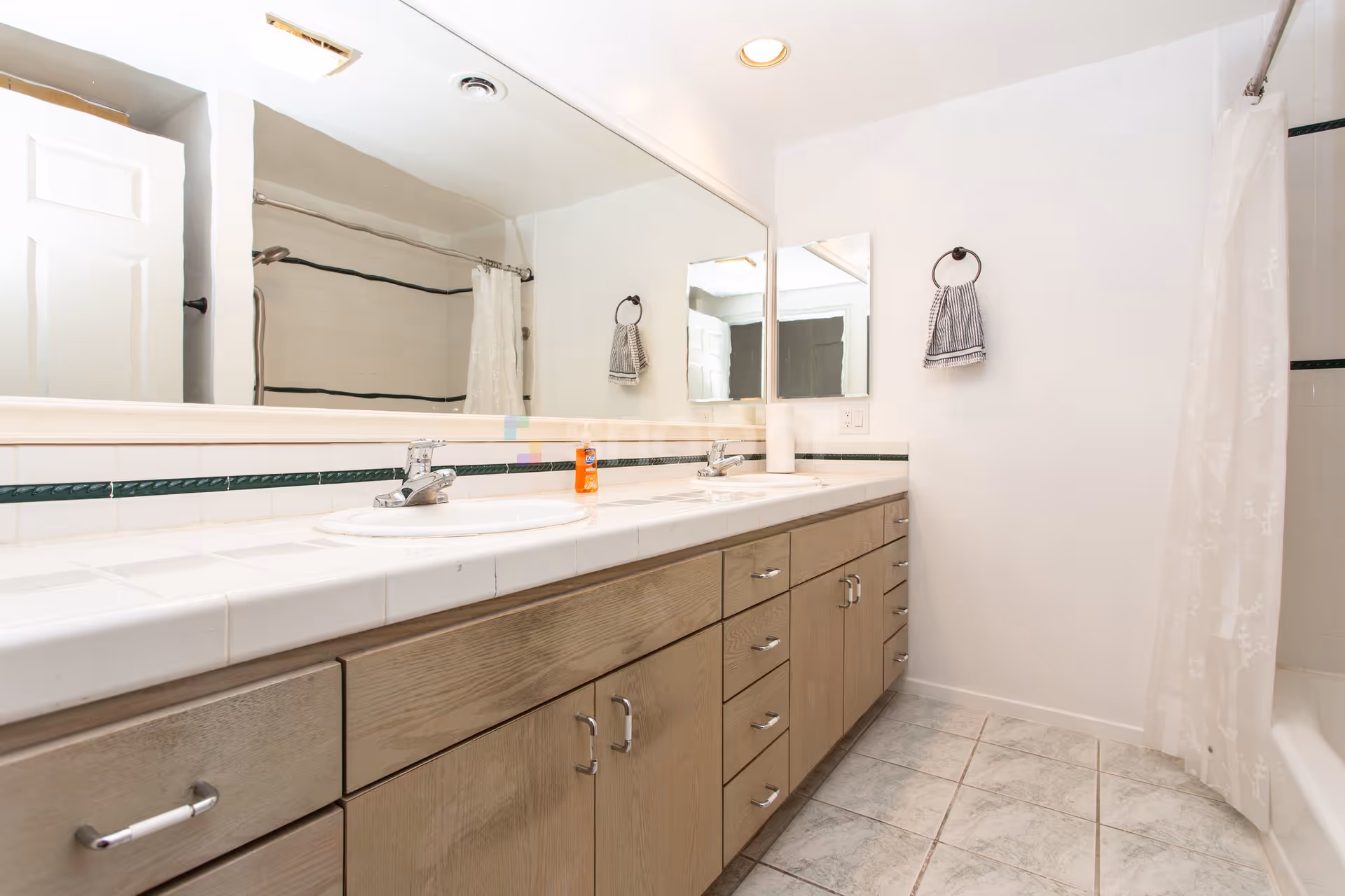 A clean bathroom with a long countertop featuring two sinks and faucets. There is a large mirror above the sinks, a hand towel hanging on a ring on the wall, and a shower with a white curtain on the right side. The floor is tiled and the cabinetry below the sinks is wooden with multiple drawers and cabinets.
