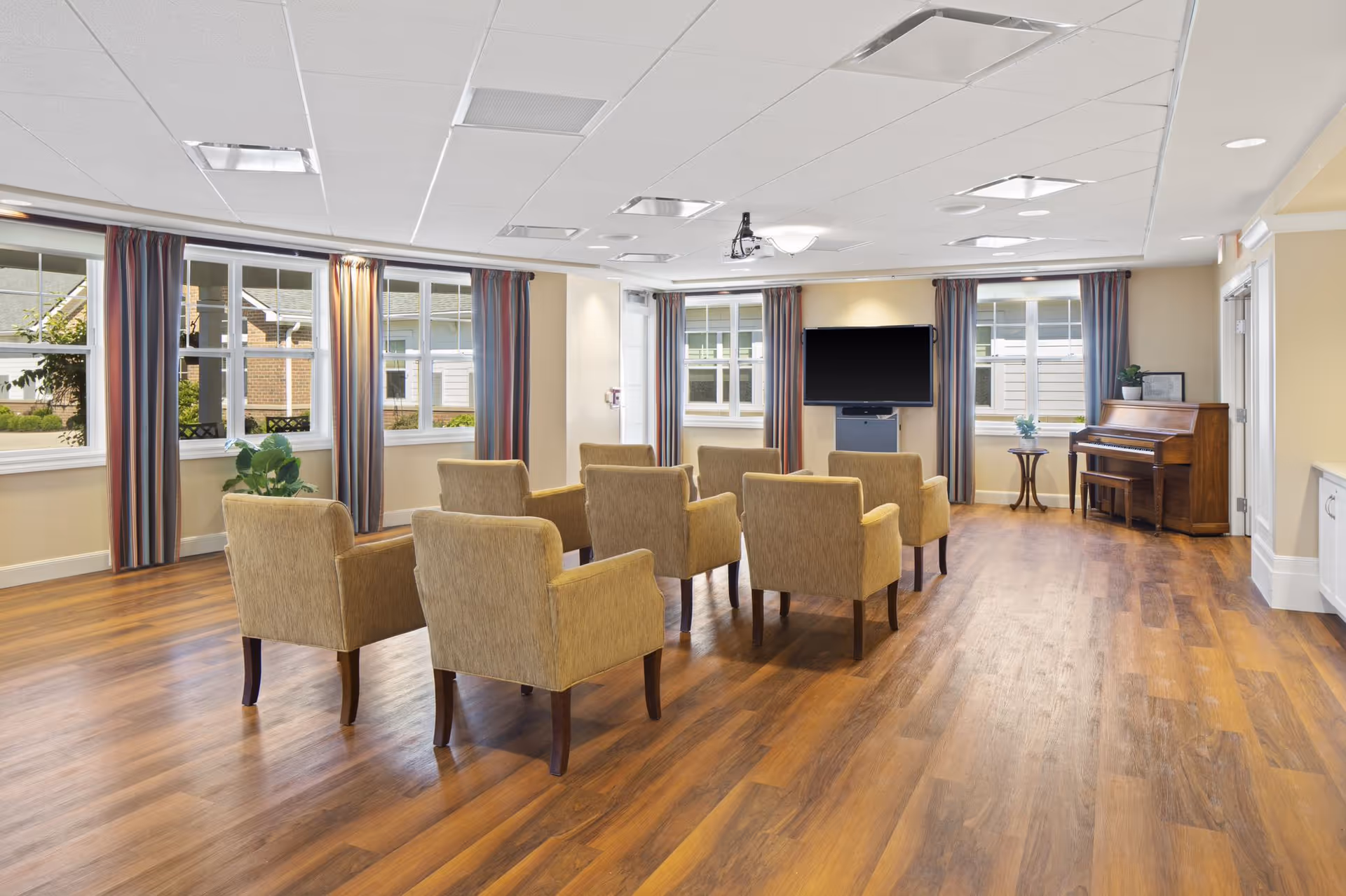 A spacious room with wooden flooring featuring two rows of beige armchairs facing a large flat-screen TV mounted on the wall. The room has multiple windows with striped curtains allowing natural light to enter. A wooden piano and a small round table with a plant are placed against the right wall. The ceiling has recessed lighting and a projector mounted in the center.