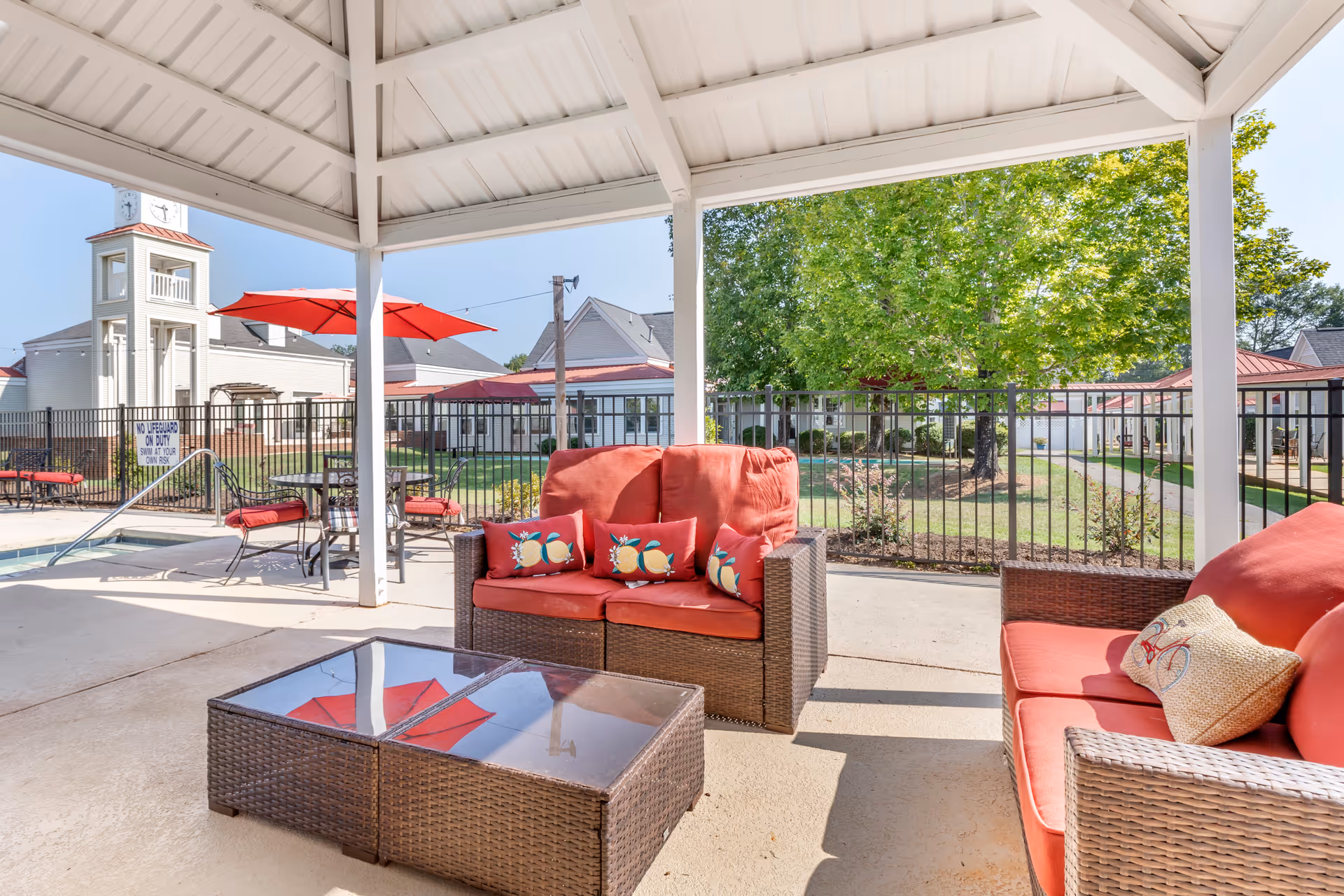 Outdoor covered seating area with wicker furniture featuring red cushions and decorative pillows. A glass-top coffee table is in front of the seating. In the background, there is a fenced pool area with a sign that reads 'No lifeguard on duty, swim at your own risk,' a table with red umbrella, and buildings with white siding and red roofs. Green trees and a clear blue sky are visible.