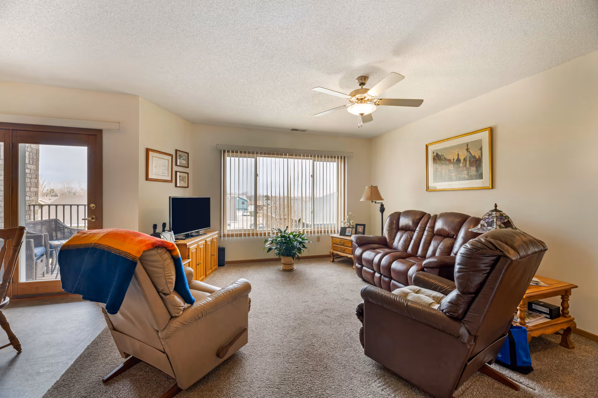 Living room with leather recliners and sofa, a TV on a wooden cabinet, ceiling fan, large window with vertical blinds, and a sliding door to a balcony.