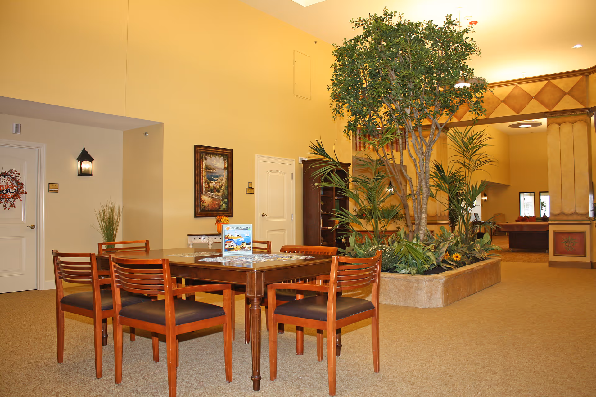 Interior view of a senior living facility common area with a wooden table and six chairs arranged around it. There is a large indoor planter with tall green plants and a tree in the background. The walls are painted light yellow, and there is a framed painting on the wall. The space has a warm and inviting atmosphere with carpeted floors and soft lighting.