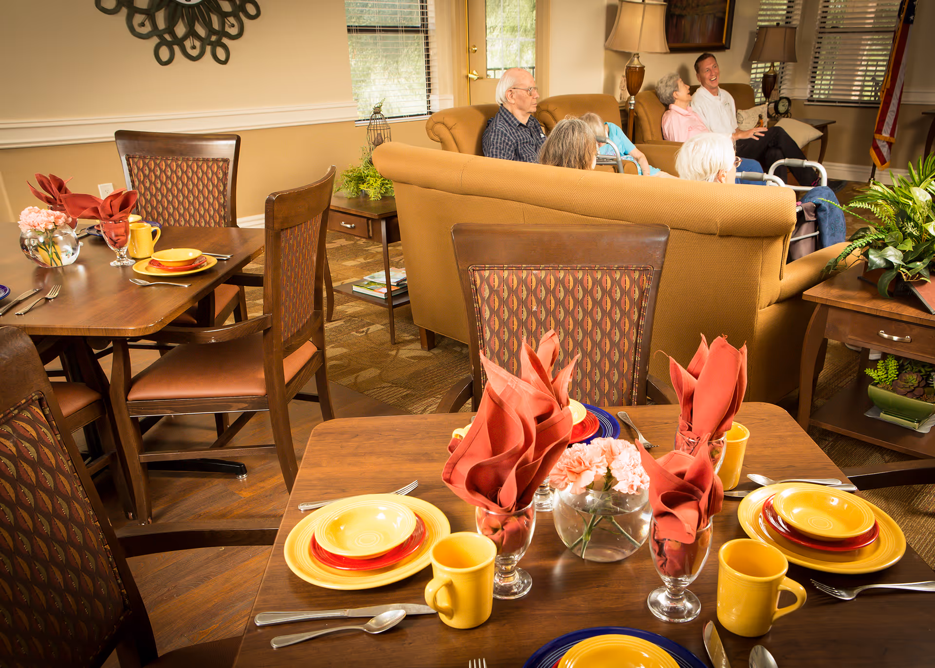 A cozy dining and living area in a senior living facility with tables set with yellow and red plates, yellow mugs, and red folded napkins in glasses. In the background, elderly residents and a caregiver are seated on a tan couch, engaged in conversation. The room is warmly lit with plants and decorative elements.