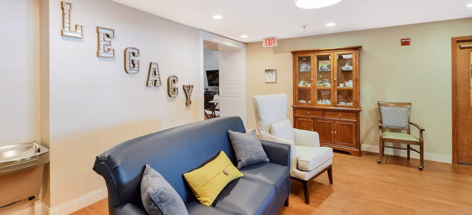 Common lounge with a blue sofa, armchairs, wooden display cabinet, and 'LEGACY' letters on the wall.