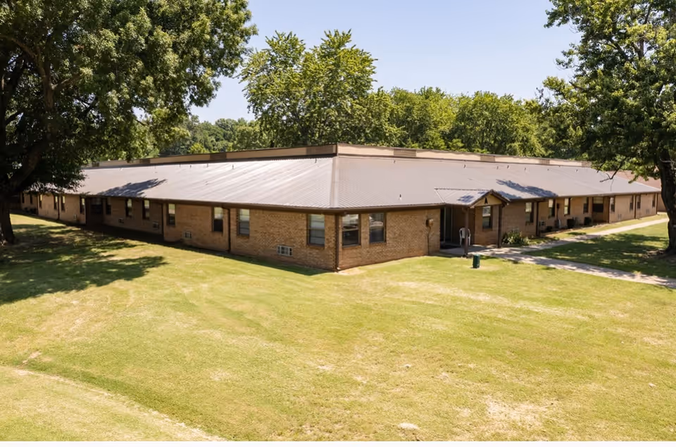 Exterior view of a single-story brick building with a metal roof surrounded by green grass and trees under a clear blue sky.