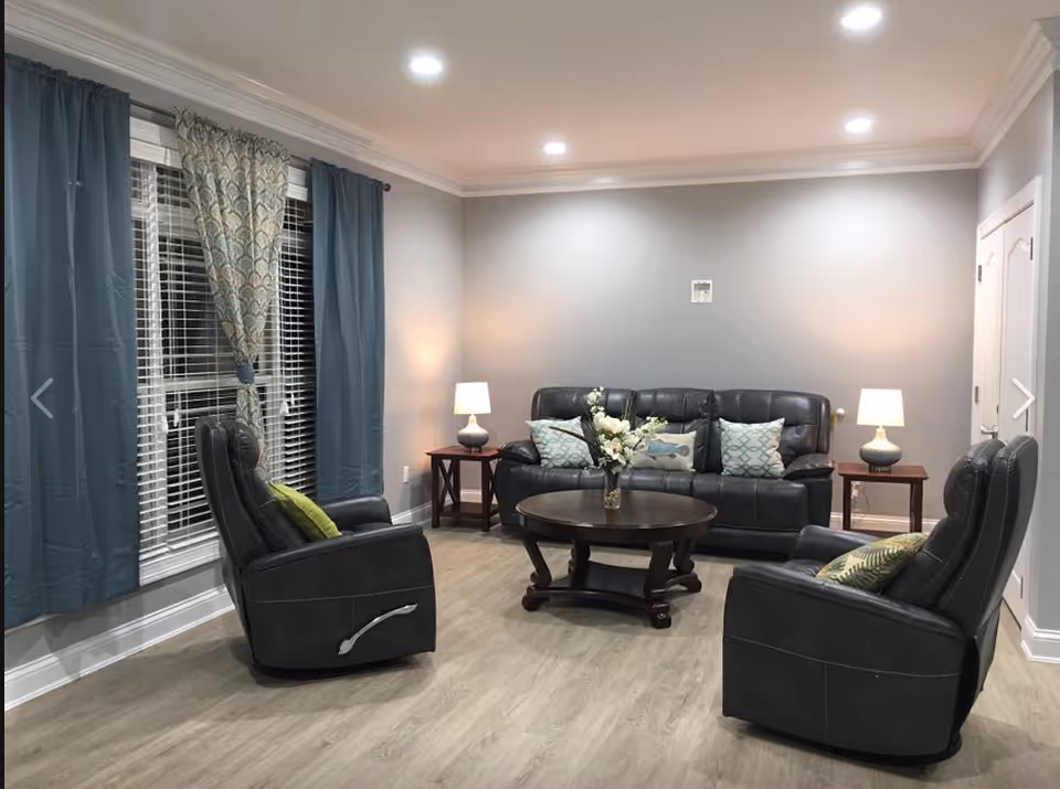 A cozy living room with a black leather sofa and two matching recliner chairs, each with decorative pillows. A round wooden coffee table with a vase of flowers sits in the center. Two side tables with lamps flank the sofa. The room has light gray walls, wood flooring, recessed ceiling lights, and large windows with blue and patterned curtains.