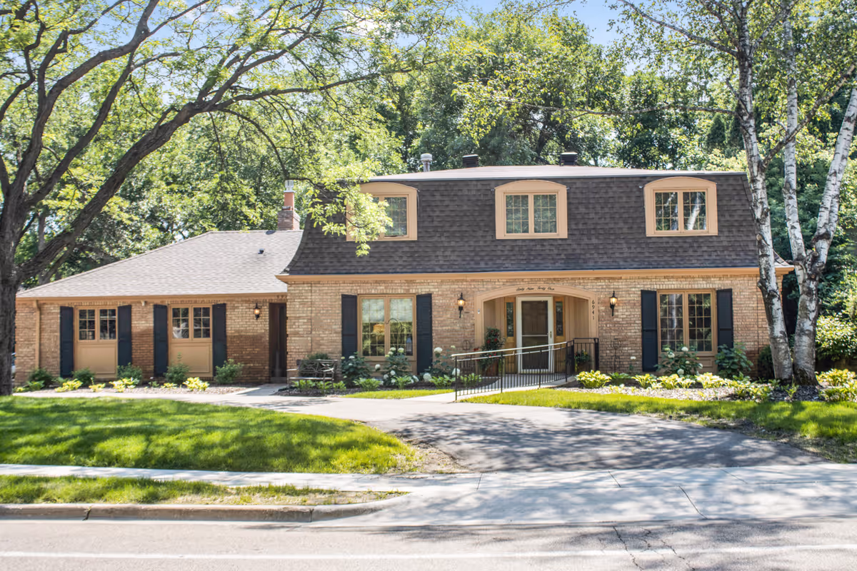 Front exterior view of a two-story brick building with a dark shingled roof, beige window frames, and black shutters. The building is surrounded by green grass, trees, and shrubs, with a paved driveway and a wheelchair ramp leading to the entrance.