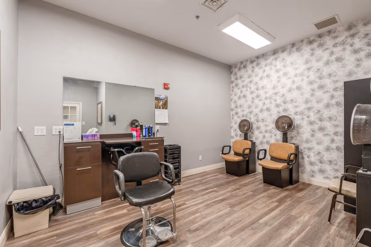 Interior view of a salon room with two brown salon chairs with hair dryers attached, a black styling chair in front of a large mirror, a cabinet with hair products, and a trash bin in the corner. The room has wood flooring and light-colored walls, one with a floral pattern.