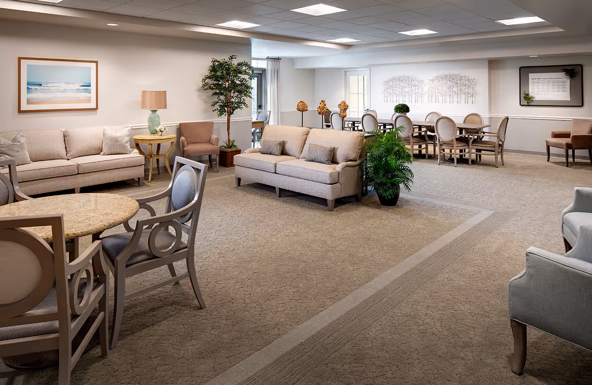 Bright, carpeted senior living common room with sofas, armchairs, round tables and a dining area in the background.