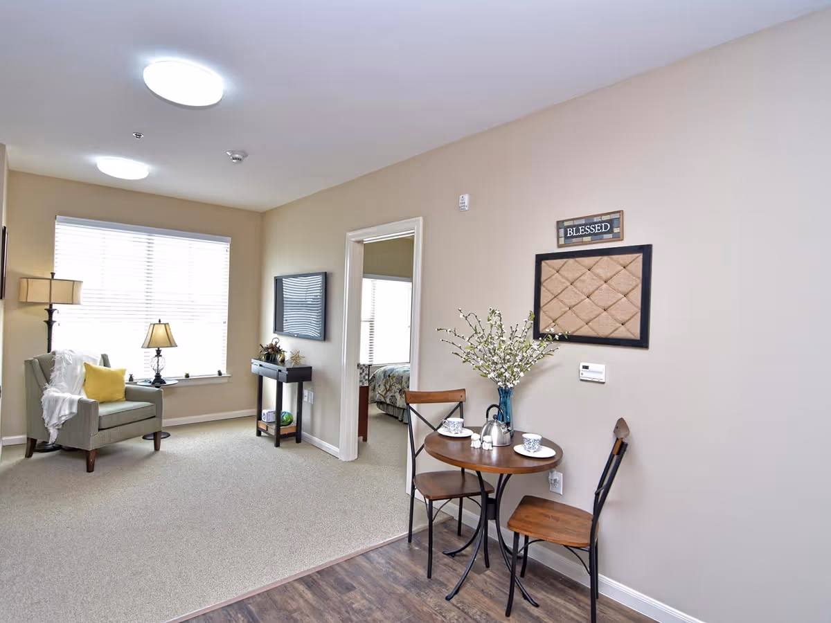 A cozy living area with a small round wooden table set for tea with two chairs, a vase with white flowers, and a wall decoration that says 'BLESSED'. To the left, there is a comfortable armchair with a yellow pillow and a white throw blanket, next to a floor lamp and a table lamp on the windowsill. A flat-screen TV is mounted on the wall above a small console table. A doorway leads to a bedroom with a bed visible through the opening. The room has beige walls and a mix of carpet and wood flooring.