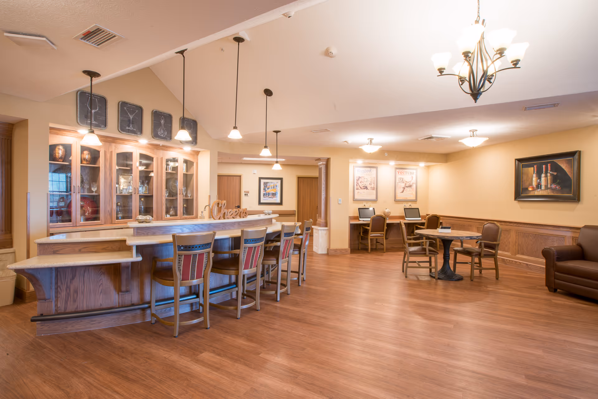 A spacious, well-lit common area with a wooden bar counter and four bar stools. Behind the bar is a glass cabinet displaying glassware. The room features wooden flooring, warm beige walls, and multiple ceiling lights including pendant lights over the bar. In the background, there are two computer desks with chairs and framed artwork on the walls. A small round table with chairs and a brown leather armchair are also visible.