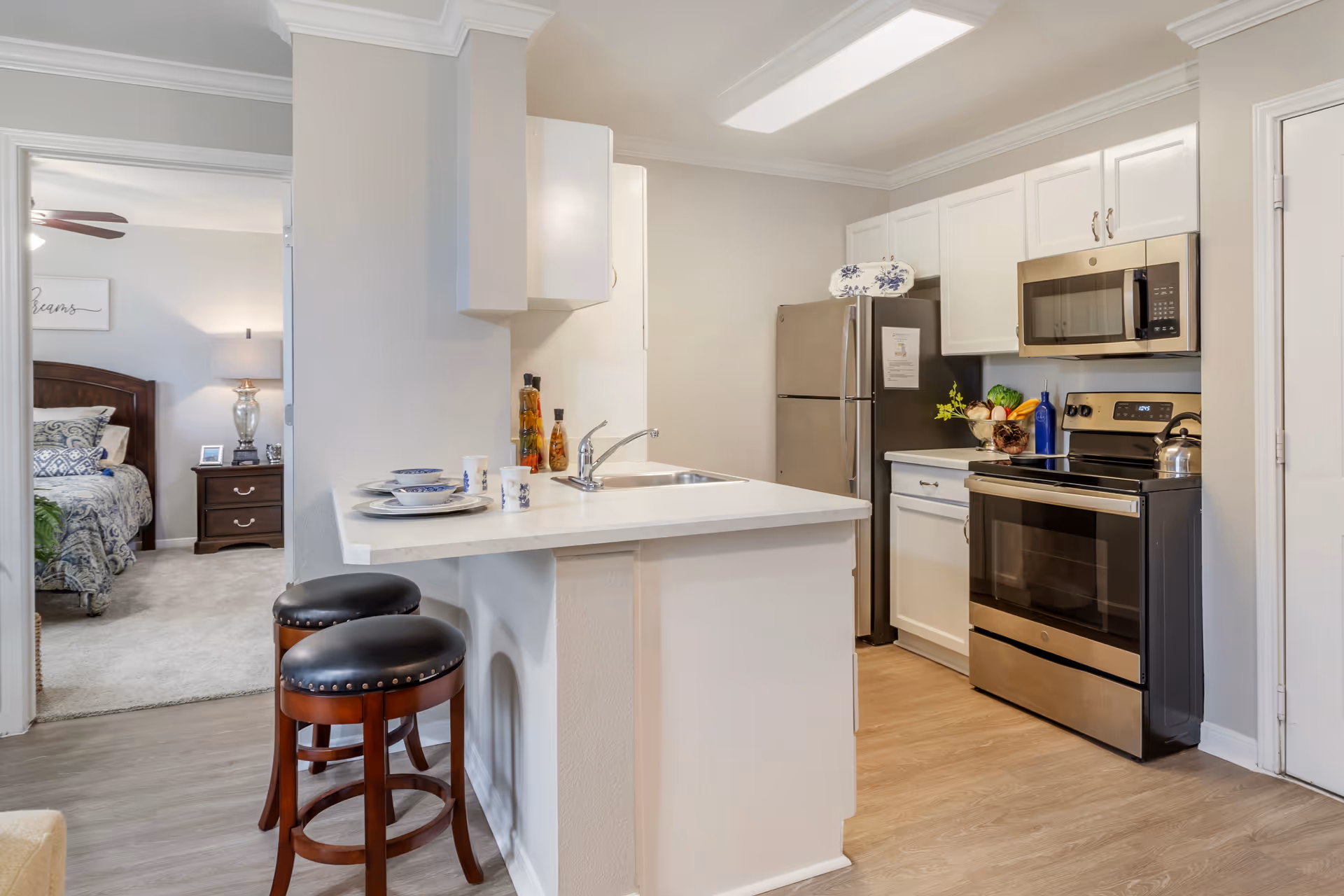 A modern kitchen with white cabinets, a stainless steel refrigerator, stove, and microwave. There is a countertop with a sink and two wooden bar stools with black cushioned seats. In the background, a bedroom with a bed, nightstand, lamp, and ceiling fan is visible through an open doorway.