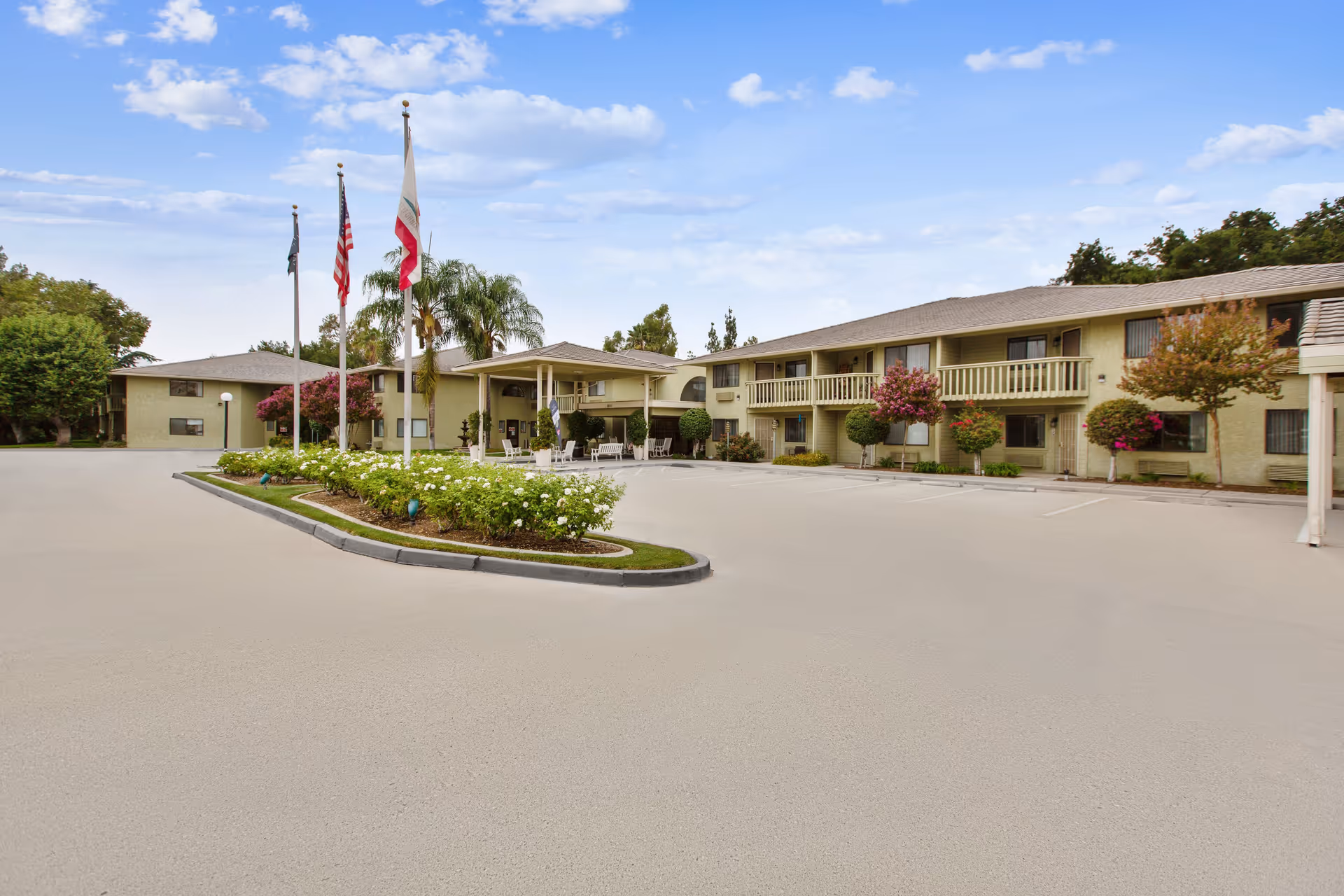 Exterior view of Truewood by Merrill, West Covina senior living facility showing a two-story building with balconies, a covered entrance, landscaped flower beds, and three flagpoles with flags against a partly cloudy sky.