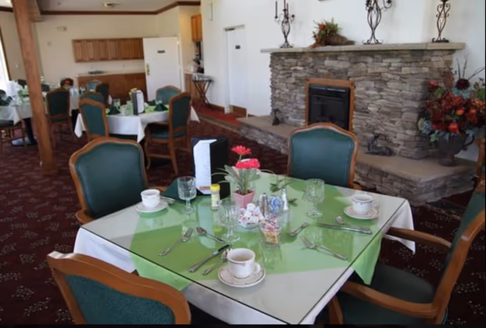 Dining room with a table set with cups, glassware and a green tablecloth, surrounded by upholstered chairs and a stone fireplace in the background.