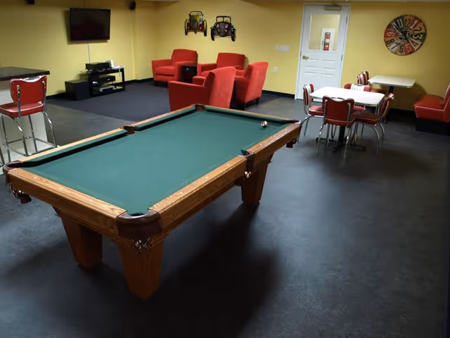 A recreational room with a green felt pool table in the center, surrounded by red cushioned chairs and a small dining area with tables and chairs. A wall-mounted TV and two decorative vintage car wall hangings are visible on the yellow walls.