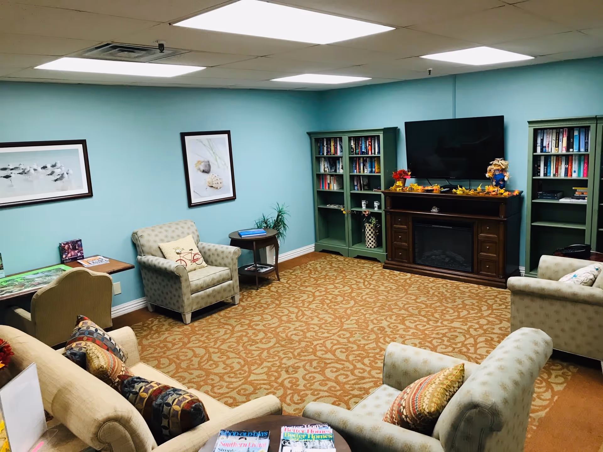 A cozy living room with light blue walls, patterned beige carpet, and several upholstered armchairs and a sofa with colorful cushions. There are two green bookshelves filled with books flanking a wooden electric fireplace with a flat-screen TV mounted above it. The room is decorated with framed pictures on the wall and small decorative items on the fireplace mantel.