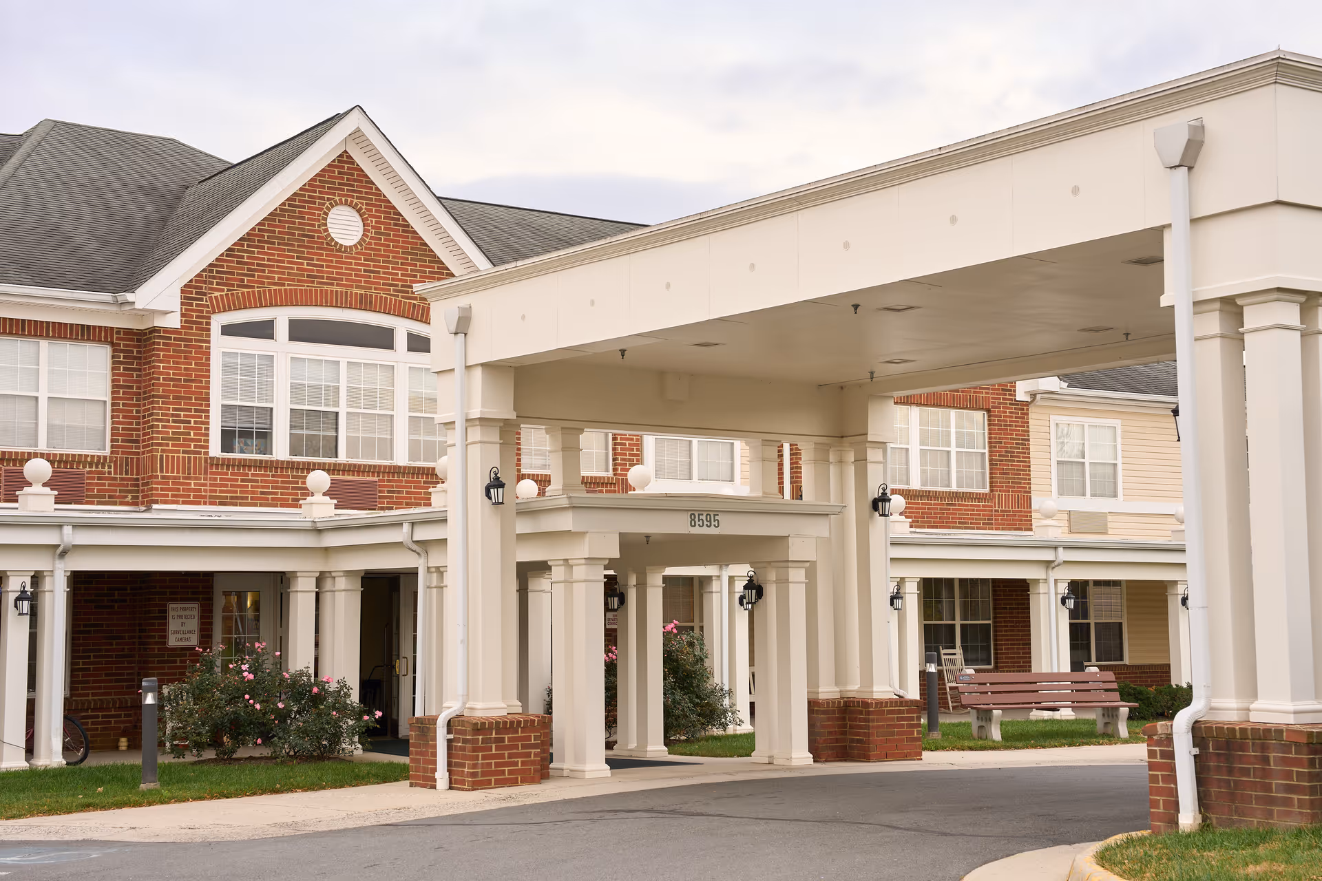 Entrance of a senior living facility with a covered drop-off area supported by white columns. The building features red brick and beige siding with multiple windows. There are benches and some landscaping with bushes and flowers near the entrance.