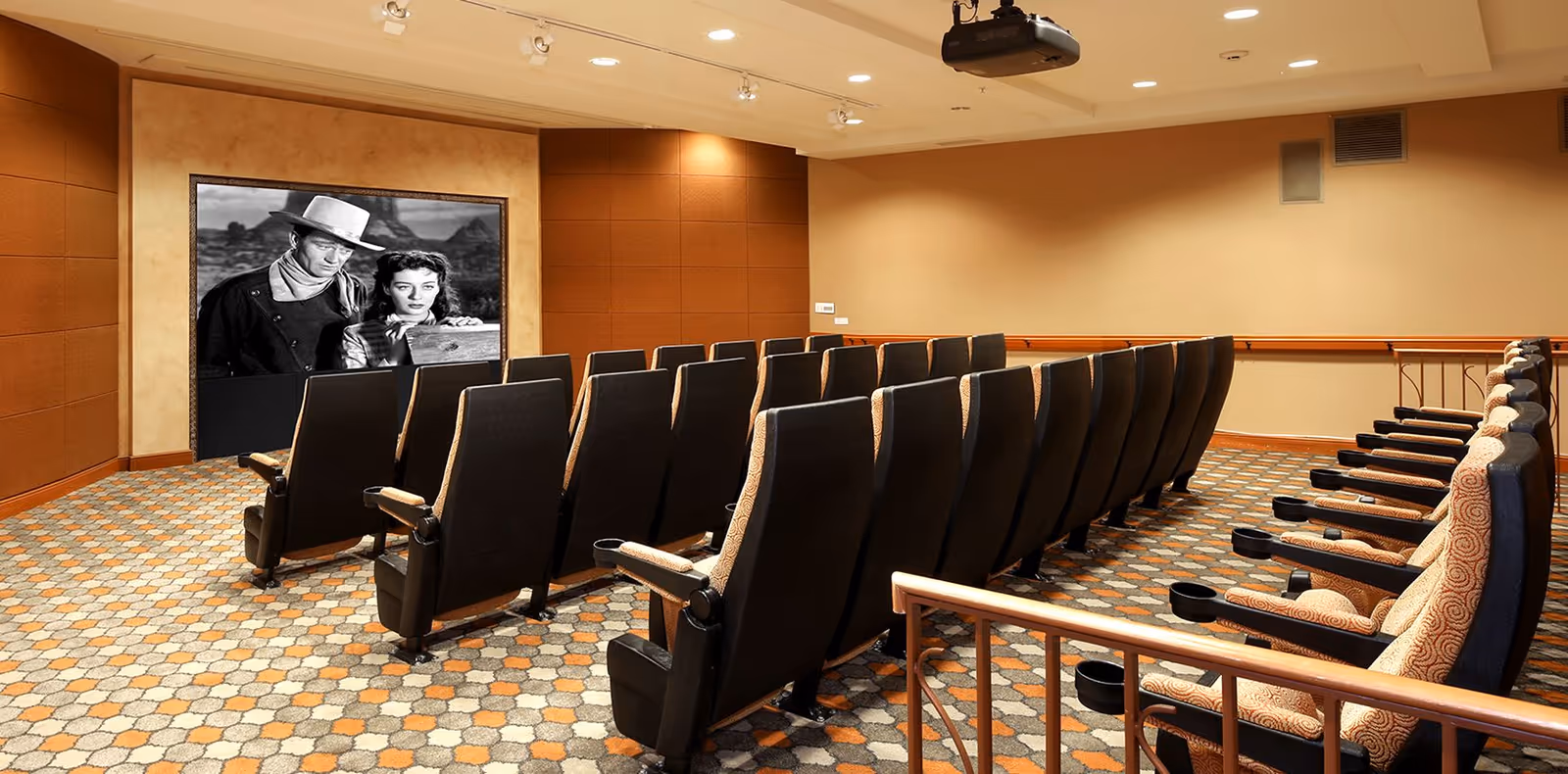 A small theater room with two rows of black cushioned seats and a side row of patterned cushioned seats, all facing a screen showing a black and white movie scene featuring a man in a cowboy hat and a woman. The room has warm beige and brown walls, a patterned carpet, and ceiling lights with a projector mounted above.