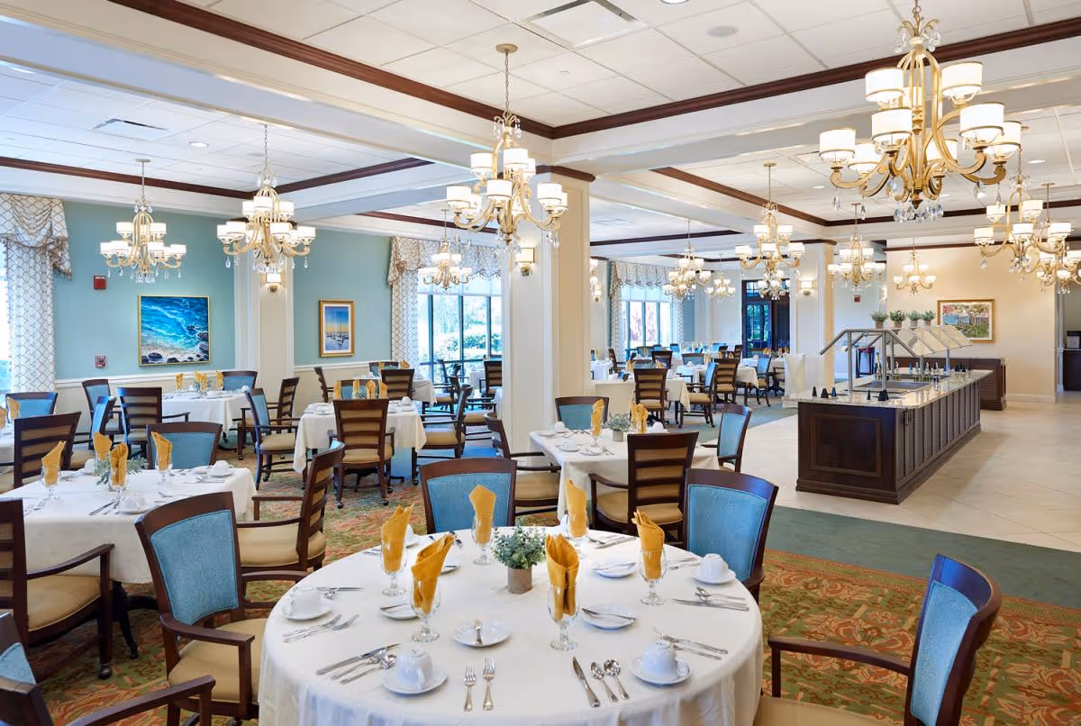 A spacious, elegant dining room with multiple round and rectangular tables set with white tablecloths, yellow folded napkins, white dishware, and silverware. The room features large windows with patterned curtains, several chandeliers hanging from the ceiling, and a buffet area with a dark wood counter. The walls are painted light blue and decorated with framed artwork.
