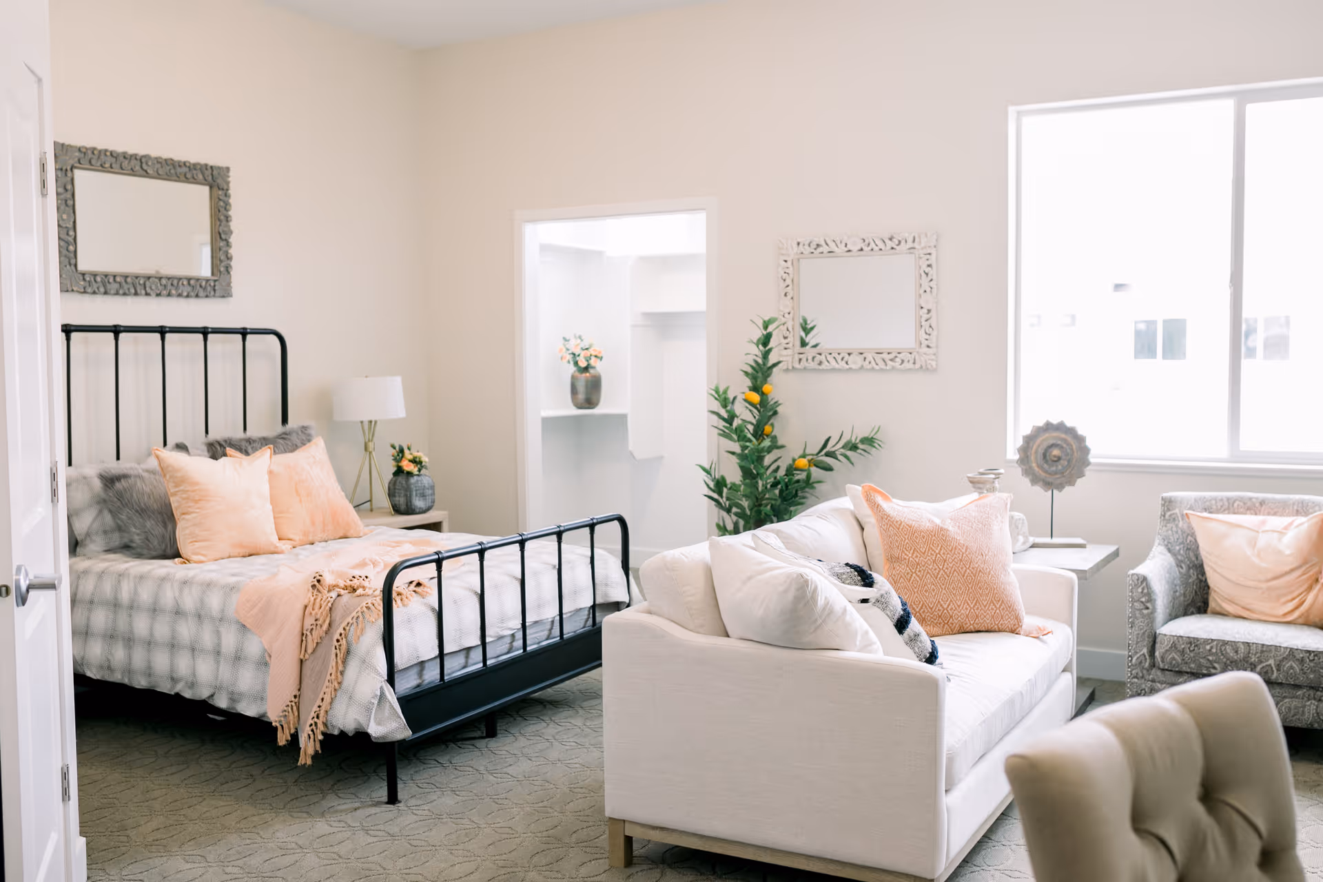 A bright and cozy room featuring a black metal bed frame with soft pillows and a throw blanket, a white sofa with decorative pillows, an armchair, a side table with a lamp, a green plant, and two decorative mirrors on the walls. A large window allows natural light to fill the space.