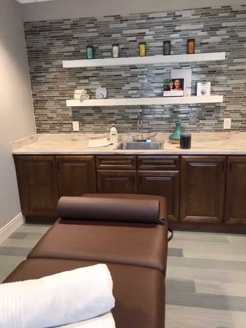 A treatment room with a brown cushioned massage or therapy table in the foreground, topped with a rolled towel. Behind it is a countertop with a sink, dark wooden cabinets, and two white floating shelves mounted on a tiled backsplash wall. The shelves hold various small items including rolled towels, candles, and decorative pieces.
