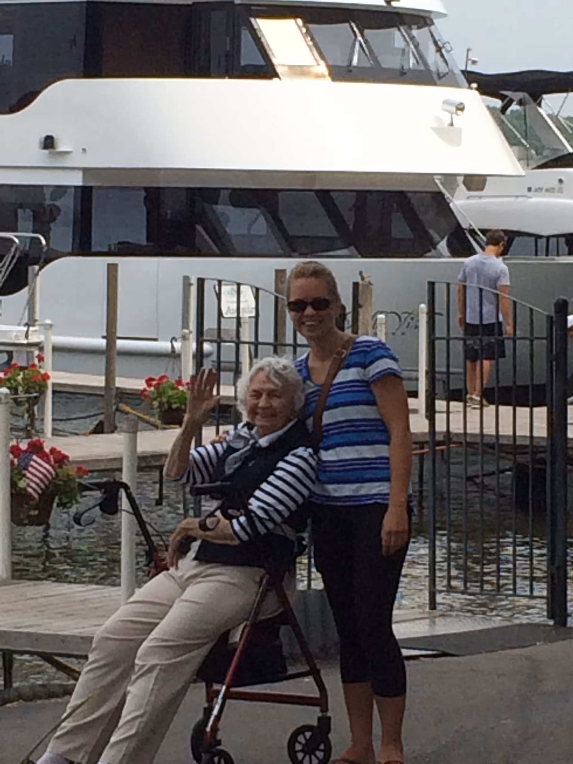 An elderly woman sitting on a walker and waving, accompanied by a younger woman standing beside her. They are outdoors near a dock with a large white boat in the background. There are flower pots with red flowers and an American flag on the dock.