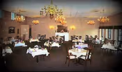 A dining room with multiple tables covered with white tablecloths and set with plates and napkins. The room features chandeliers hanging from the ceiling, a fireplace in the background, and large windows on one side allowing natural light to enter.