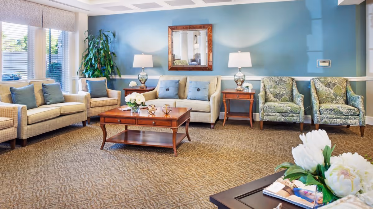 A bright and inviting living room with beige sofas and patterned armchairs arranged around a wooden coffee table. The walls are painted blue with a large mirror hanging above the sofa. Two table lamps with white shades sit on wooden side tables. A large window with blinds lets in natural light, and there are green plants and white flowers adding a touch of nature to the room.