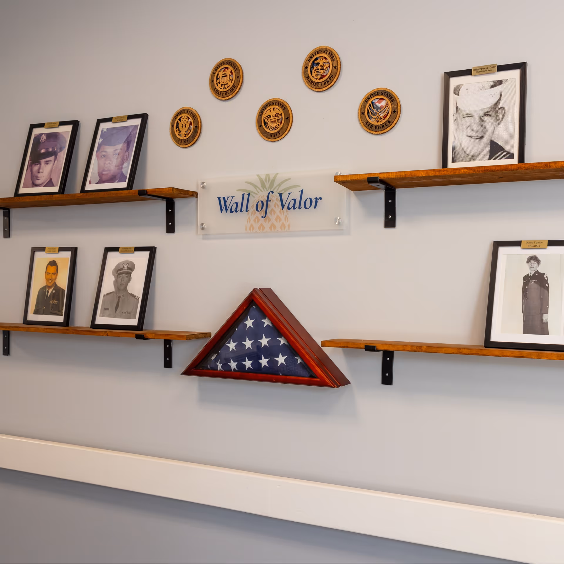 A wall display titled 'Wall of Valor' featuring framed black and white photographs of military personnel, military branch emblems, and a folded American flag in a triangular wooden case mounted on wooden shelves against a light gray wall.