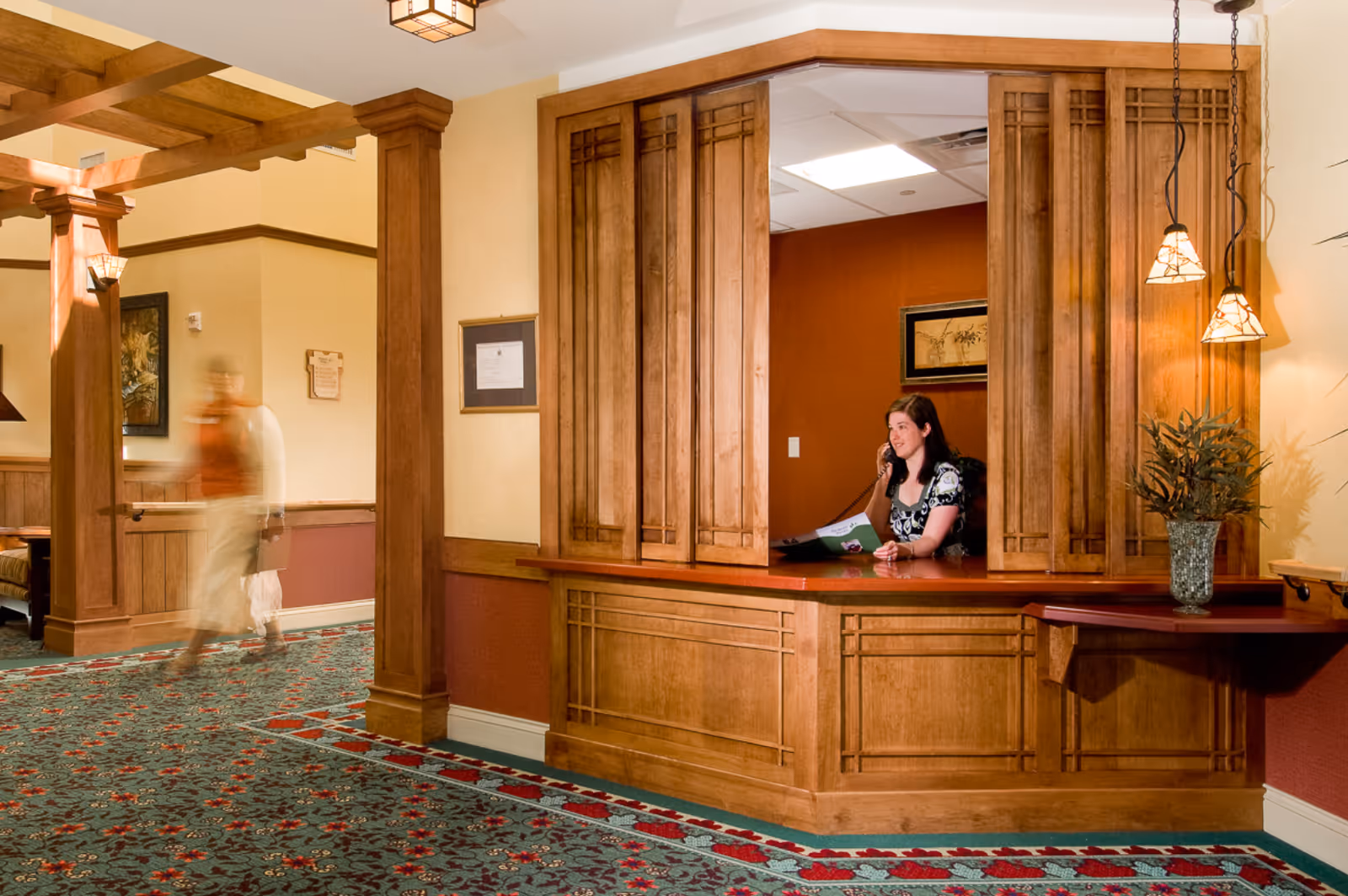 Reception area in a senior living facility with a woman sitting behind a wooden reception desk talking on the phone. The area features warm wood paneling, decorative hanging lights, a patterned carpet, and a blurred figure walking in the hallway.