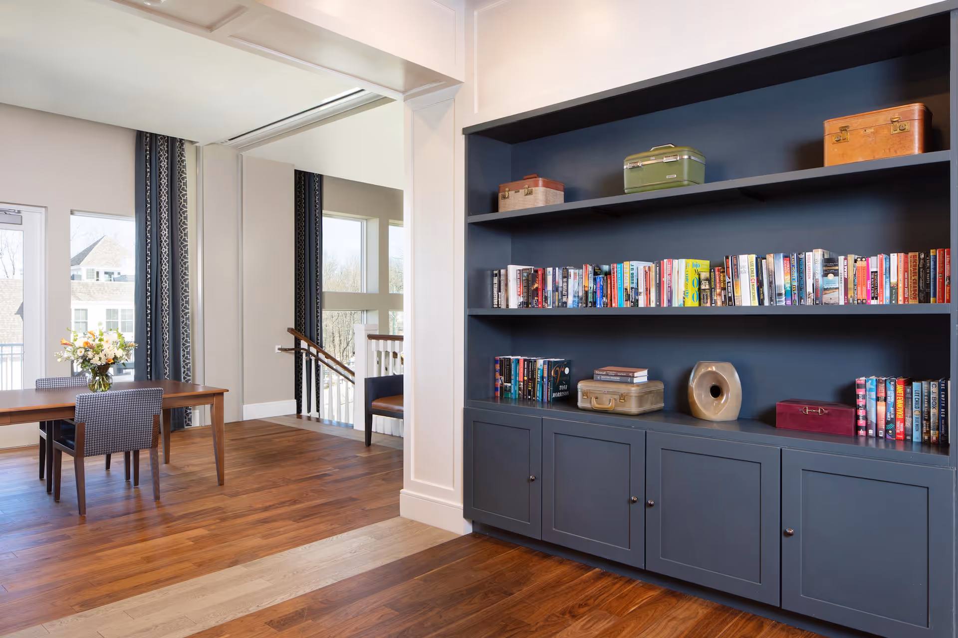 Interior view of a senior living facility showing a built-in dark blue bookshelf filled with books and decorative items. To the left, there is a wooden table with two chairs and a vase of flowers, with large windows and a door letting in natural light. The floor is wooden and the walls are painted in neutral tones.