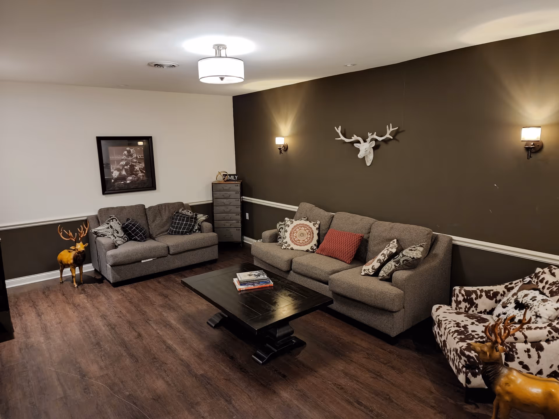 A cozy living room with dark wood flooring, a gray three-seater sofa with decorative pillows, a matching loveseat, and a patterned armchair. A dark wooden coffee table with books sits in the center. The walls are painted half white and half dark brown, with a white deer head decoration mounted on the brown wall and a framed black-and-white photo on the white wall. Two small deer statues are placed near the furniture, and the room is softly lit by ceiling and wall lights.