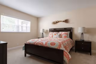 Bright bedroom with a dark wood bed dressed in pink-and-white patterned bedding, two bedside tables with lamps, and a window letting in natural light.