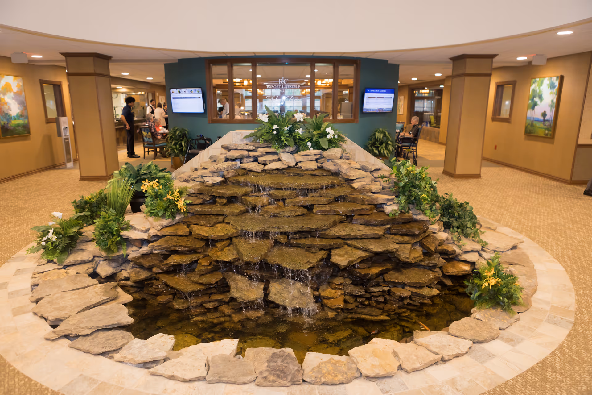 Indoor water feature with cascading rocks and surrounding plants in the center of a spacious, well-lit common area with carpeted floors, beige walls, and framed artwork. People are visible in the background near seating areas and a reception or dining area.