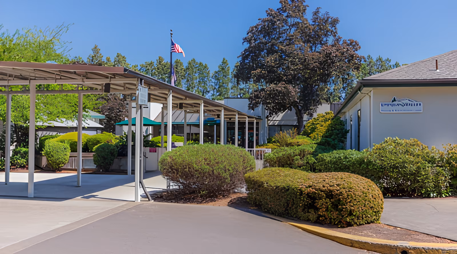 Covered entrance and walkway to a nursing facility with landscaped shrubs, an American flag, and a building sign.