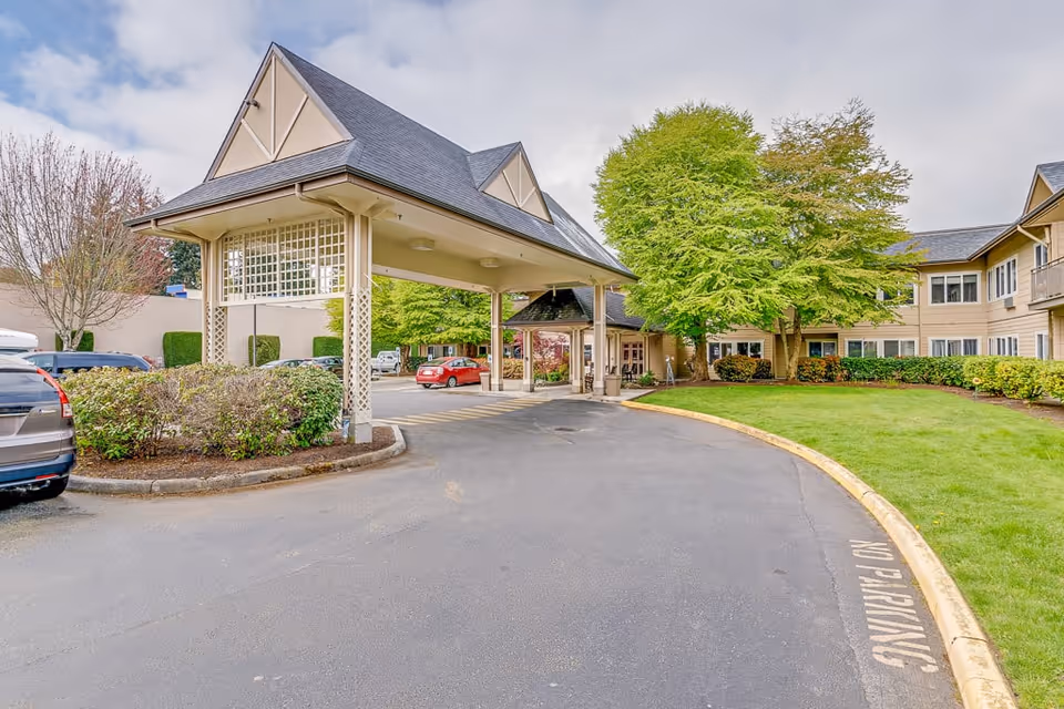 Exterior view of Solstice Senior Living At Normandy Park showing the driveway with a covered entrance, parked cars, green trees, and the building facade under a partly cloudy sky.