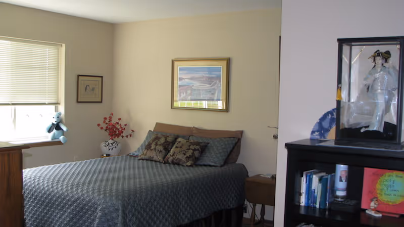 A bedroom with a neatly made bed covered with a patterned gray bedspread and several pillows. There is a window with closed blinds on the left wall, a small teddy bear sitting on the window sill, and framed artwork hanging on the walls. A vase with red flowers is placed on a small table next to the bed. On the right side, there is a dark wooden bookshelf with books and decorative items, including a doll in a glass case on top.